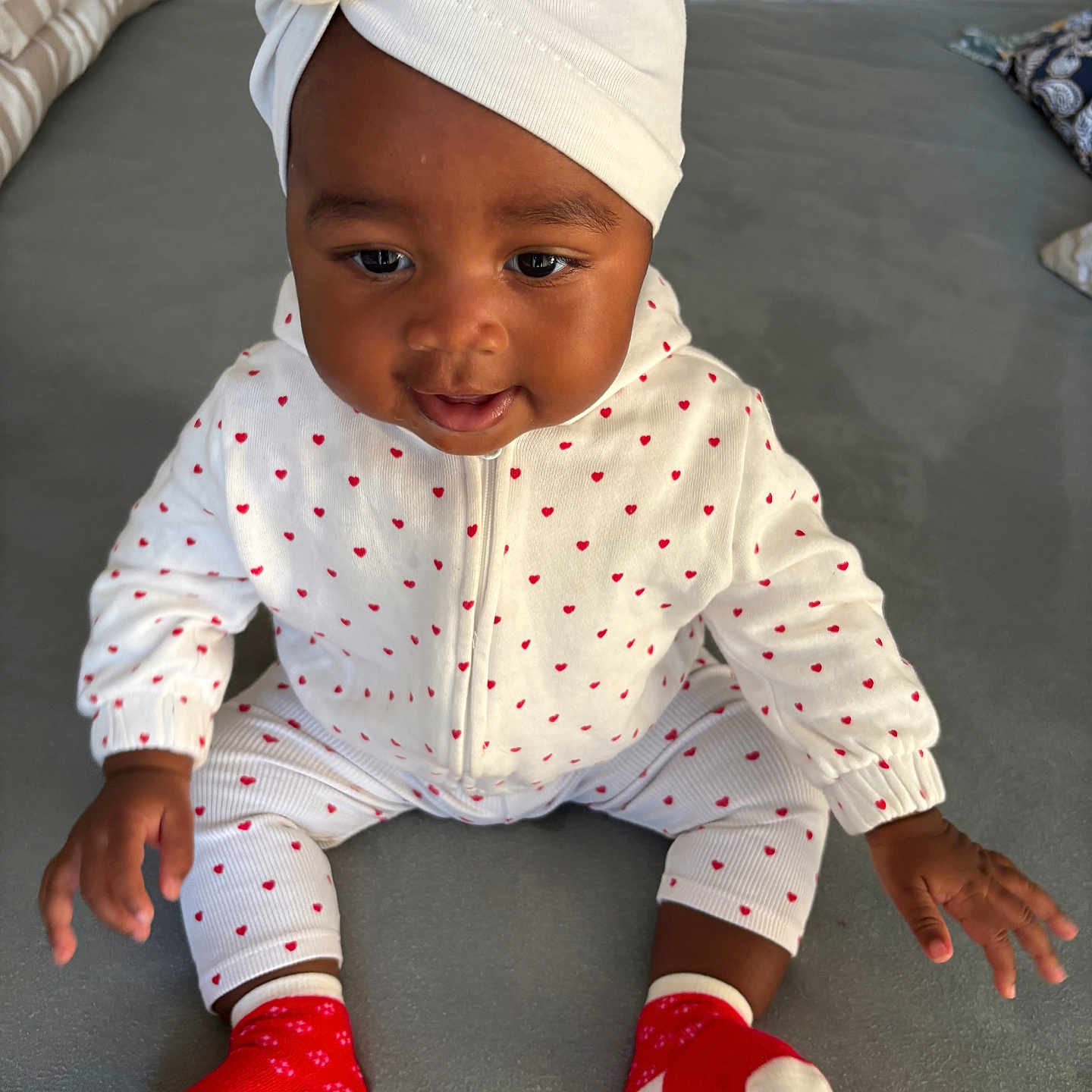 Imany participe au concours pour gagner de l'argent avec cette photo : baby, child, clothing, cozy, cute, feet, gray_surface, hands, headwrap, indoor, infant, person, portrait, red_hearts, red_socks, sitting, smiling, turban, white_outfit, young_child