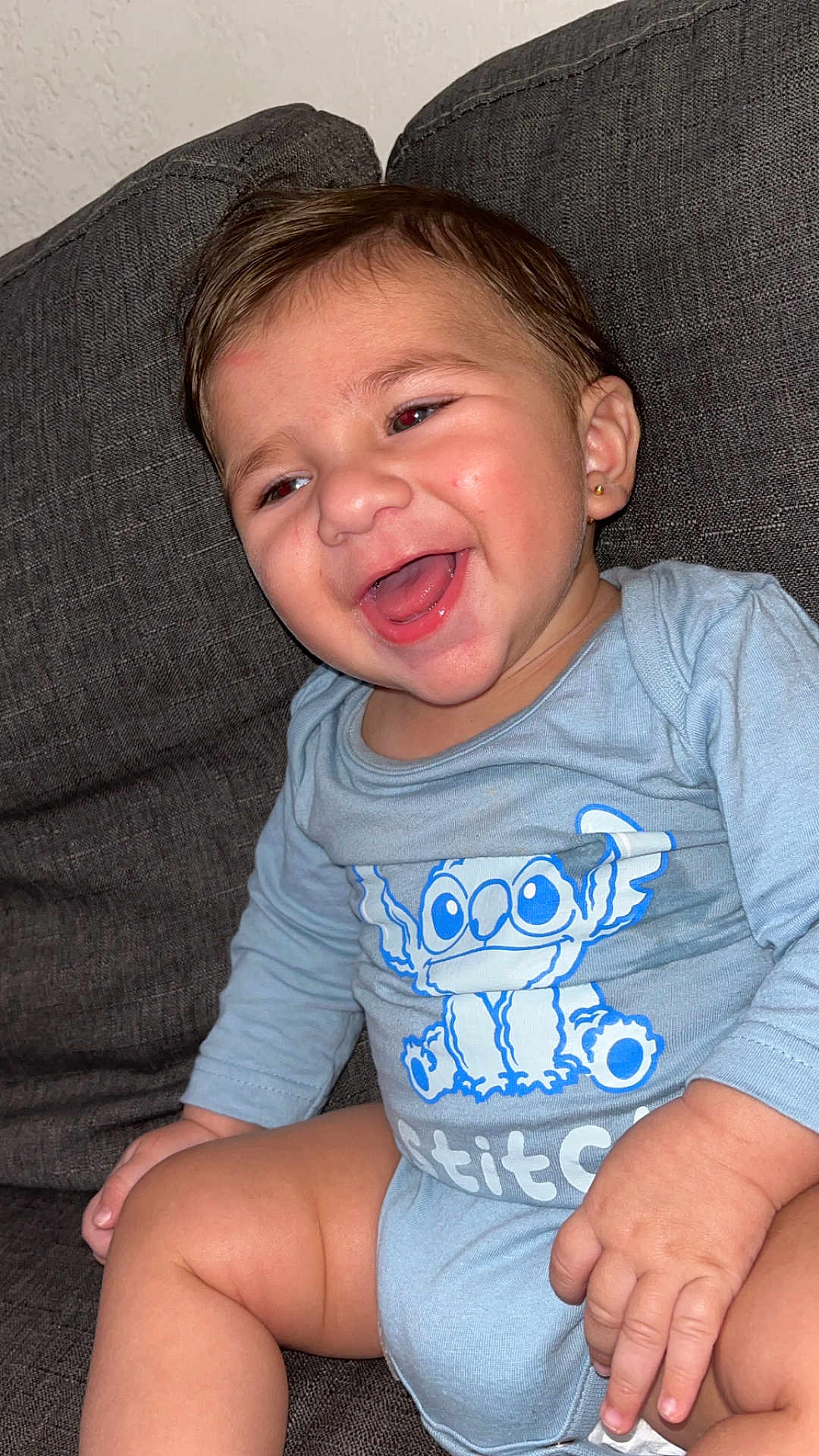 Julian participe au concours pour gagner de l'argent avec cette photo : baby, child, smiling, laughing, blue_onesie, stitch, sitting, couch, indoor, happy, cute, person, infant, young_child, skin, hair, earring, hand, leg, clothing