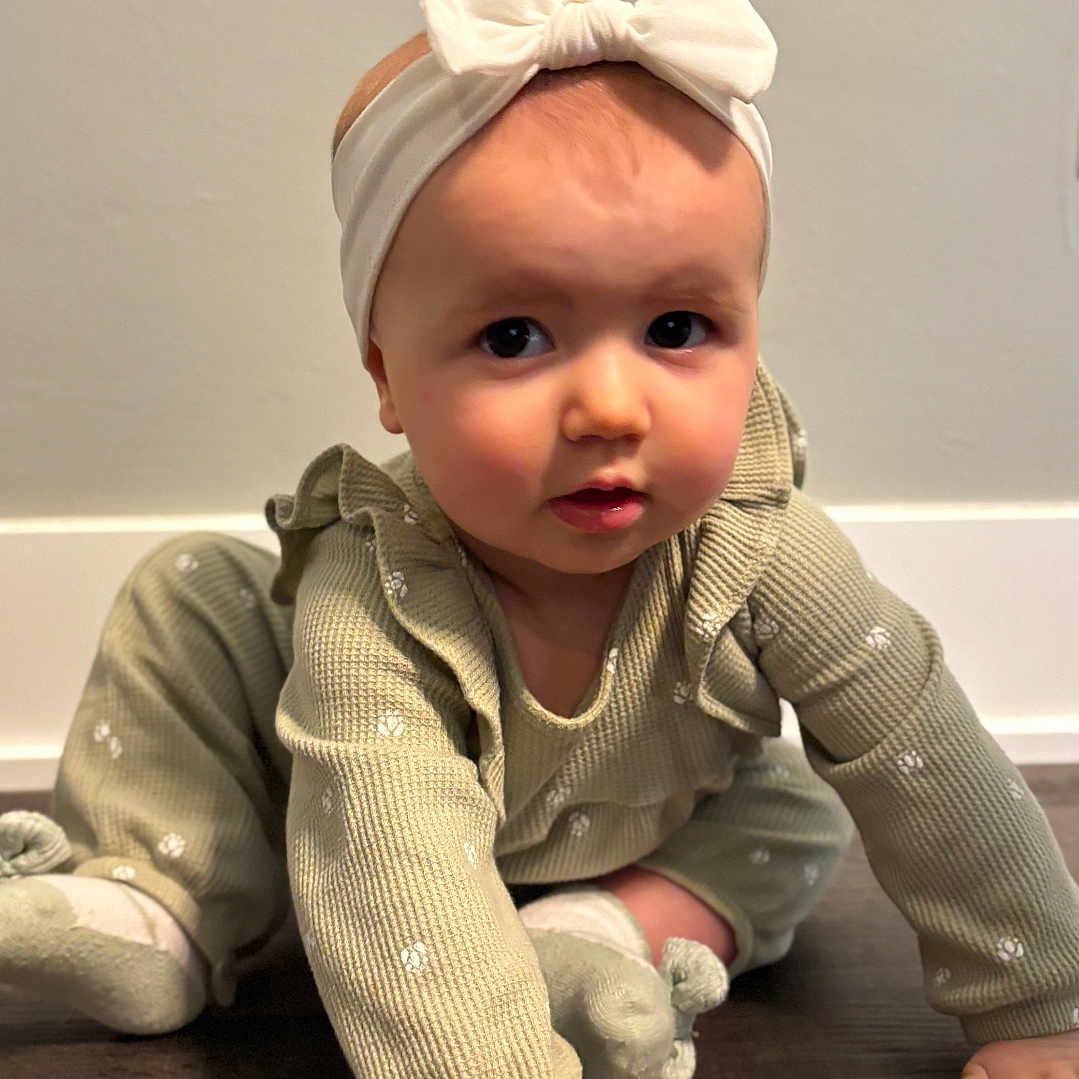Polina is registered to the contest to win money with this photo: baby, bow, child, crawling, curious, cute, exploring, face, floor, green_outfit, hand, headband, indoor, infant, long_sleeves, person, portrait, socks, wooden_floor, young_child