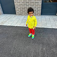 Mateo participe au concours pour gagner de l'argent avec cette photo : child, toddler, yellow_shirt, red_pants, colorful_sneakers, pacifier, brick_wall, door, pavement, outdoor, standing, person, curly_hair, urban, casual_clothing, footwear, sidewalk, young_child, daylight, street