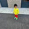 child, toddler, yellow_shirt, red_pants, colorful_sneakers, pacifier, brick_wall, door, pavement, outdoor, standing, person, curly_hair, urban, casual_clothing, footwear, sidewalk, young_child, daylight, street