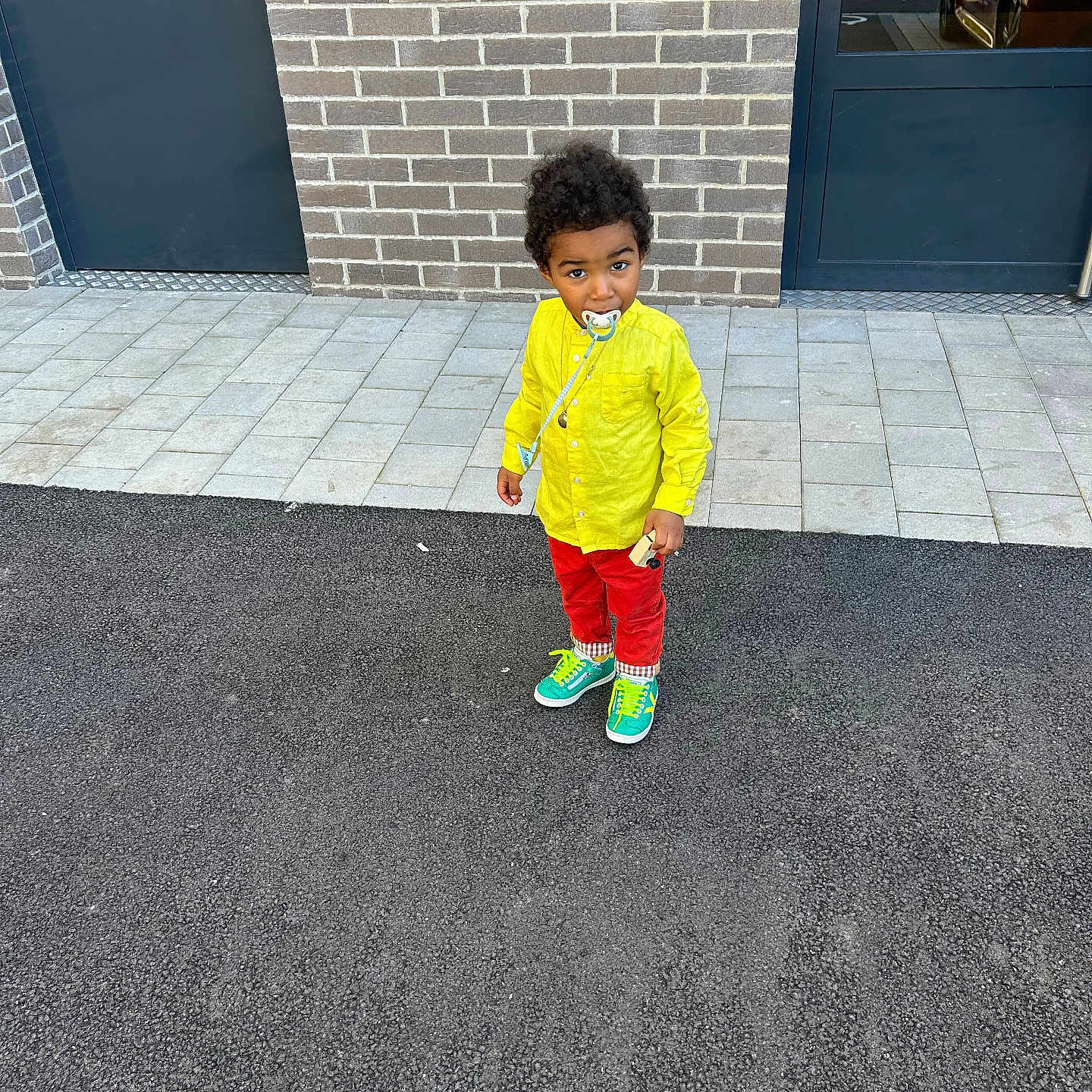 Mateo participe au concours pour gagner de l'argent avec cette photo : child, toddler, yellow_shirt, red_pants, colorful_sneakers, pacifier, brick_wall, door, pavement, outdoor, standing, person, curly_hair, urban, casual_clothing, footwear, sidewalk, young_child, daylight, street