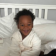 Kartier is registered to the contest to win money with this photo: toddler, child, smiling, robe, bed, pillow, blanket, curly_hair, indoor, happy, person, white_clothing, cozy, furniture, head, face, sitting, cute, home, relaxed