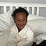 toddler, child, smiling, robe, bed, pillow, blanket, curly_hair, indoor, happy, person, white_clothing, cozy, furniture, head, face, sitting, cute, home, relaxed
