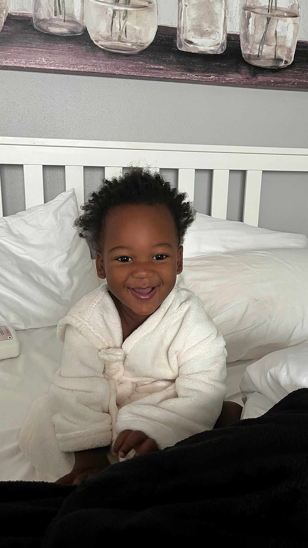 Kartier is registered to the contest to win money with this photo: toddler, child, smiling, robe, bed, pillow, blanket, curly_hair, indoor, happy, person, white_clothing, cozy, furniture, head, face, sitting, cute, home, relaxed