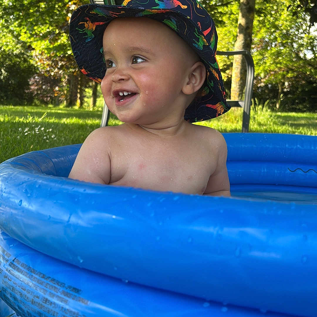 Kobe is registered to the contest to win money with this photo: toddler, child, pool, inflatable_pool, hat, dinosaur_hat, outdoor, grass, trees, sunlight, smiling, water, summer, happy, play, nature, baby, bare_chest, blue, green