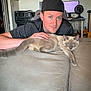 man, cat, siamese_cat, couch, indoor, pet, computer, printer, fan, black_cap, relaxed, home_office, furniture, person, animal, light_skin, blue_eyes, casual, technology, hand