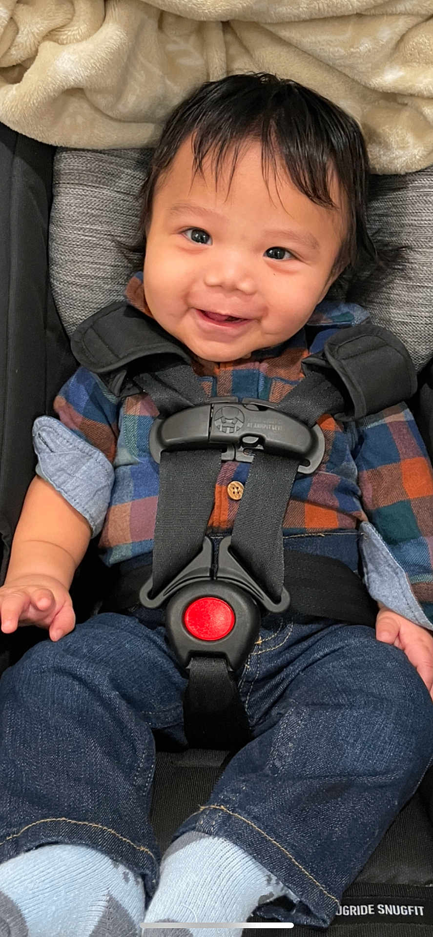 Silas is registered to the contest to win money with this photo: child, baby, infant, smiling, car_seat, safety_harness, buckle, red_button, plaid_shirt, flannel_shirt, jeans, socks, blanket, hair, face, eyes, cheeks, portrait, close_up, cute