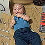 baby, infant, child, lying_down, tools, wrenches, screwdriver, cardboard, stroller, wheel, license_plate, grease_smudges, blue_shirt, navy_pants, barefoot, playful_expression, surprised_face, toolbox, floor, home_workshop