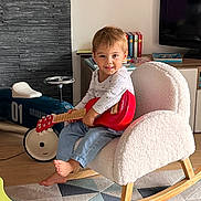 Robin participe au concours pour gagner de l'argent avec cette photo : child, toddler, rocking_chair, toy_guitar, indoor, living_room, car_toy, books, furniture, floor_rug, barefoot, smiling, wooden_floor, white_chair, casual_clothing, playful, colorful, home, cute, person