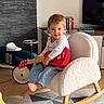 child, toddler, rocking_chair, toy_guitar, indoor, living_room, car_toy, books, furniture, floor_rug, barefoot, smiling, wooden_floor, white_chair, casual_clothing, playful, colorful, home, cute, person