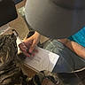 Ozzy is registered to the contest to win money with this photo: arm, blue_shirt, cap, casual, cat, cozy, curious, distraction, floor, furniture, glass_table, hand, indoor, notebook, paw, pen, person, reflection, tabby_cat, writing