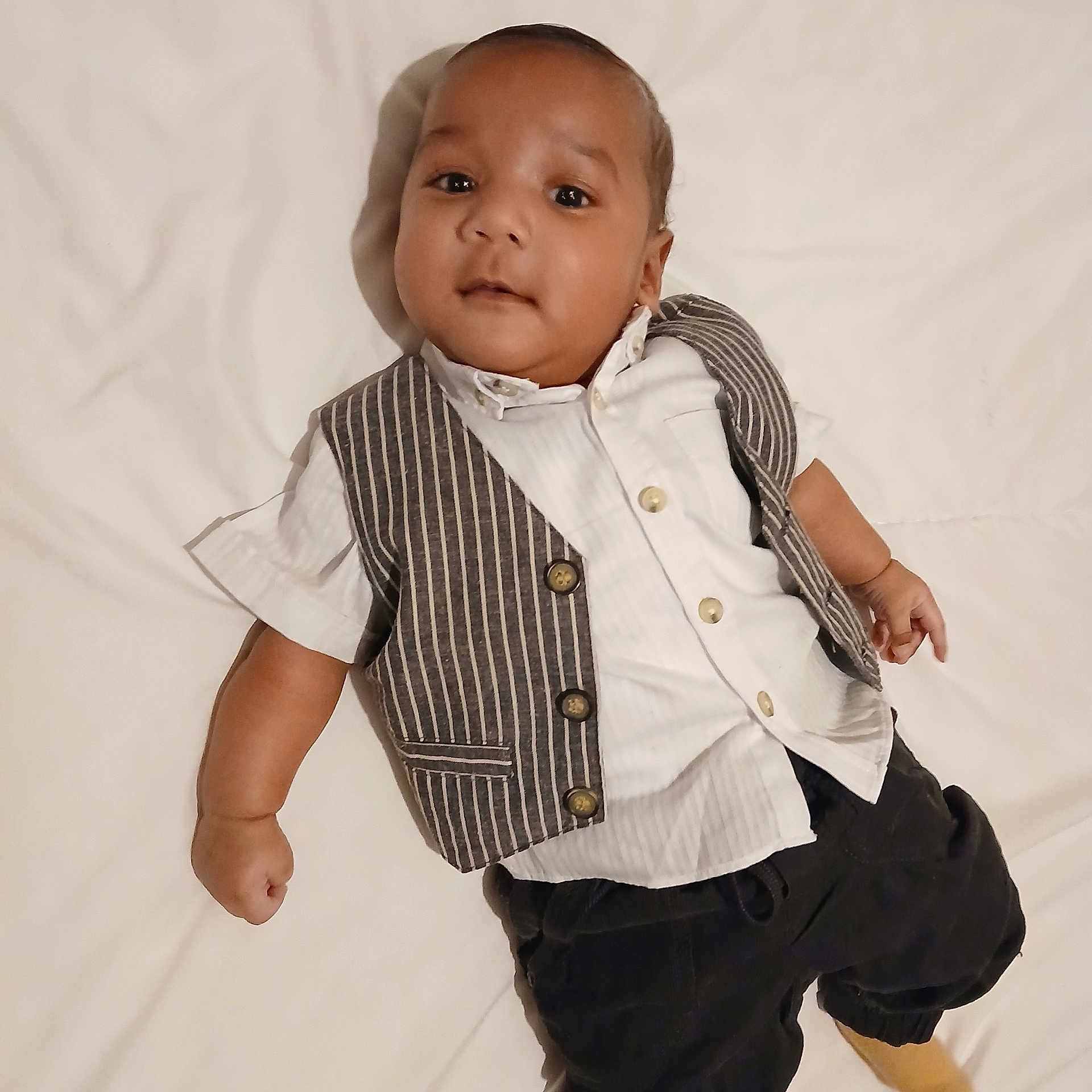 Peni is registered to the contest to win money with this photo: baby, casual_clothing, child, comfortable, cute, face, footwear, hand, indoors, infant, looking_at_camera, lying_down, pants, portrait, shirt, small_person, soft_surface, vest, white_background, young_child