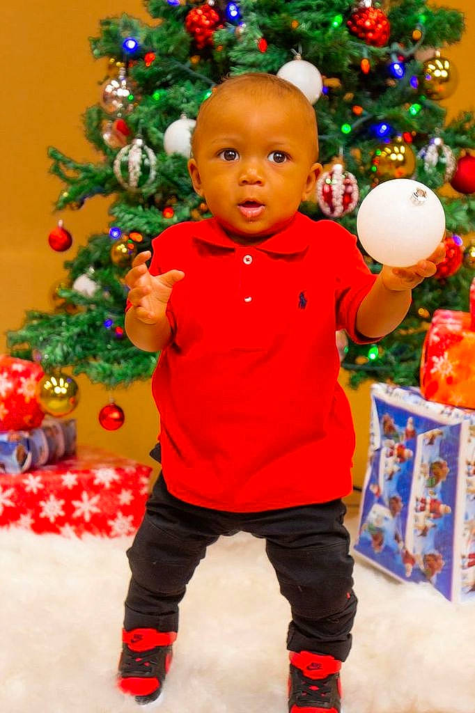Jamal is registered to the contest to win money with this photo: baby_toddler_clothing, christmas, christmas_decoration, christmas_ornament, christmas_tree, facial_expression, fun, green, happy, holiday_ornament, human_body, ornament, people, people_in_nature, person, plant, sleeve, standing, toddler, tree