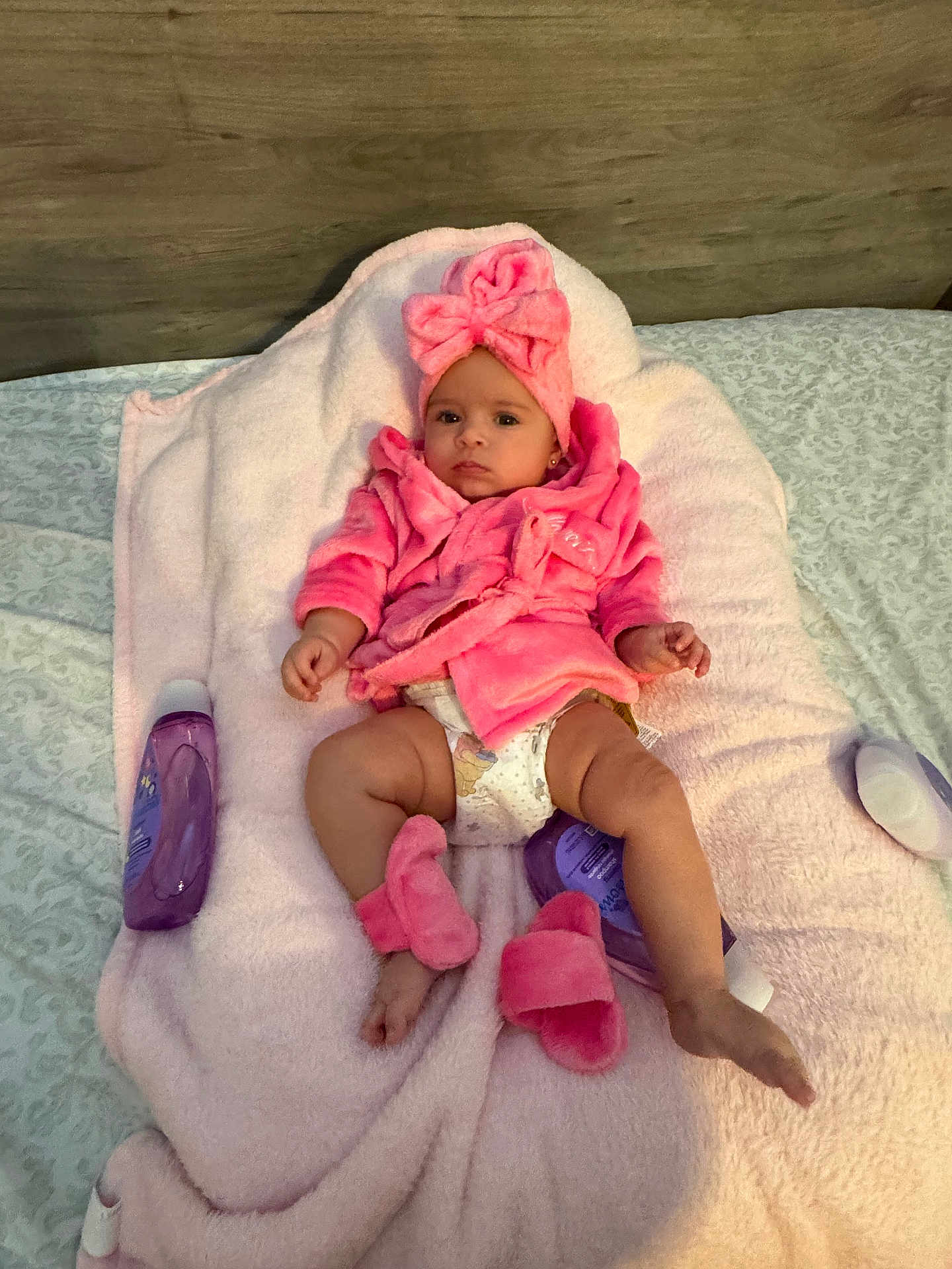 Samantha is registered to the contest to win money with this photo: baby, pink, robe, headwrap, blanket, lotion, slippers, diaper, bed, wooden_headboard, cute, infant, soft_texture, relaxed, indoors, child, person, skin, clothing, laying_down