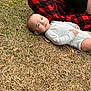 baby, infant, onesie, grass, outdoor, person, hand, plaid_pants, red_and_black, side_eye, skeptical_expression, dry_grass, casual_clothing, nature, candid, child, young_child, human, relaxed, daytime