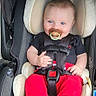 baby, baby_accessory, baby_girl_or_boy, baby_toddler, black_shirt, blue_eyes, car_seat, child, child_safety, clothing, cute, footwear_free, indoors, infant, pacifier, portrait, red_pants, safety, seatbelt, smiling