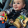 Atlas is registered to the contest to win money with this photo: baby, child, car_seat, toy, pacifier, plaid_shirt, denim, smile, face, hand, clothing, seatbelt, infant, cute, indoors, person, bright_colors, closeup, young, happy