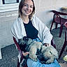 animal, building, casual, chair, daylight, dog, friendship, fur, happy, jeans, outdoor, pavement, pet, plastic_chair, poster, relaxed, seated, smile, window, woman