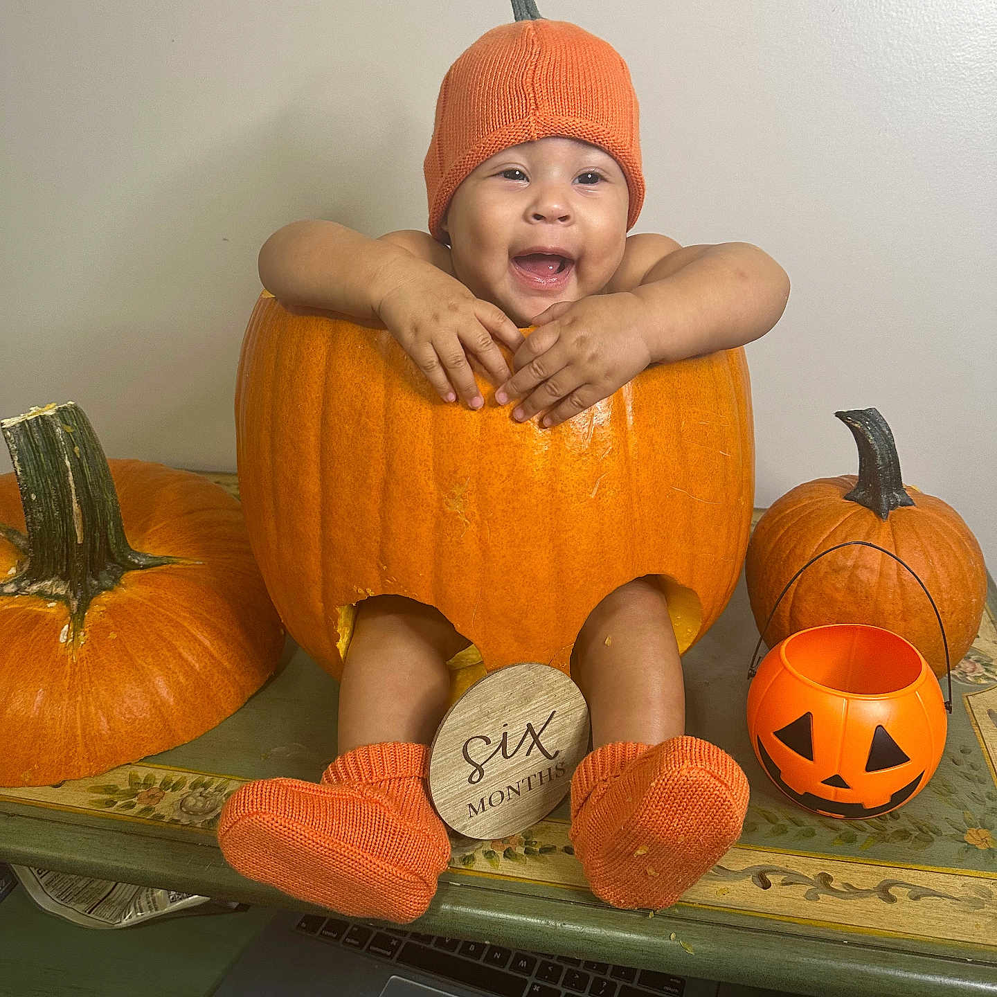 Arya is registered to the contest to win money with this photo: baby, pumpkin, orange, hat, socks, table, carved_pumpkin, smiling, six_months, decor, halloween, child, festive, cute, indoor, person, holiday, seasonal, pumpkin_bucket, happy