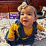Kyrah joined the competition — help win amazing prizes! baby, smiling, headband, yellow_clothing, denim_dress, blanket, bed, indoor, furniture, dresser, television, happy, child, person, cute, portrait, sitting, clothing, room, cozy