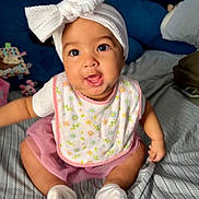 Kyrah is registered to the contest to win money with this photo: baby, infant, child, smiling, headband, bow, bib, pink_skirt, socks, bed, pillow, toy, patterned_bedspread, cute, happy, sitting, indoors, portrait, face, person