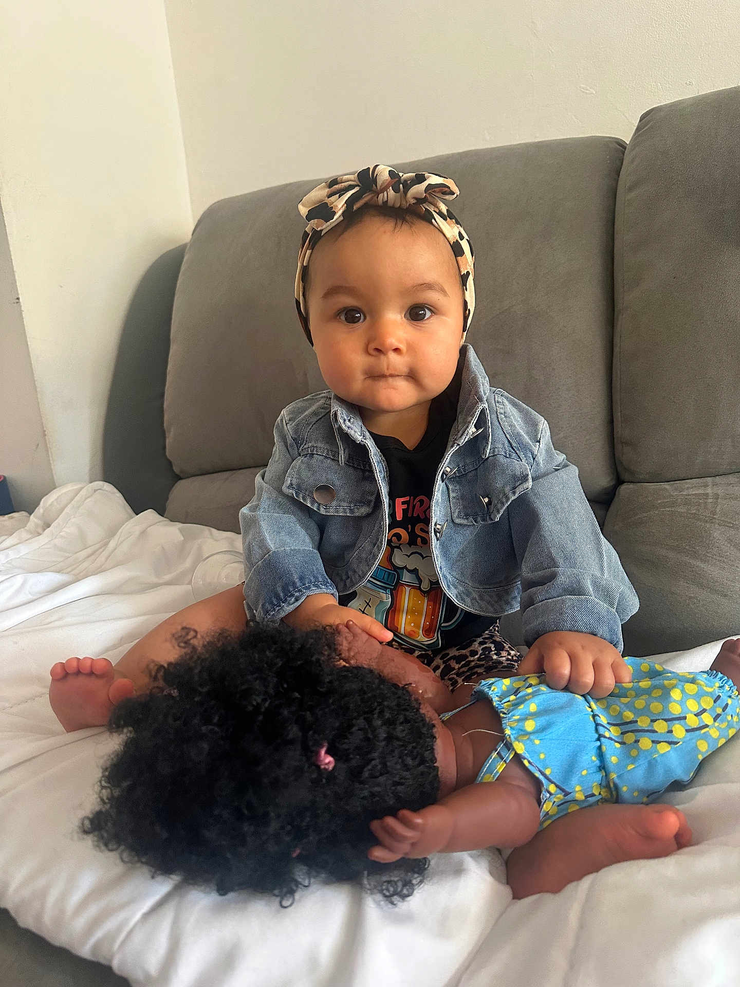 Aïna a rejoint le concours — aidez-le/la à gagner de superbes lots ! baby, child, doll, toy, headband, denim_jacket, curly_hair, blue_dress, polka_dot, couch, indoor, white_bedspread, serious_expression, skin, person, infant, cute, holding, sitting, portrait