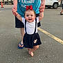 Annabelle joined the competition — help win amazing prizes! baby, child, toddler, red_bow, white_shirt, suspenders, black_skirt, barefoot, adult_hands, parking_lot, car, suv, traffic_cones, blue_shirt, flip_flops, outdoor, asphalt, person, walking, support
