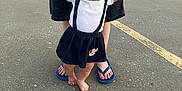 Annabelle is registered to the contest to win money with this photo: baby, child, person, holding_hands, parking_lot, red_bow, white_shirt, black_skirt, adult, blue_shirt, flip_flops, car, sidewalk, outdoor, building, store_sign, footwear, cute, walking, support