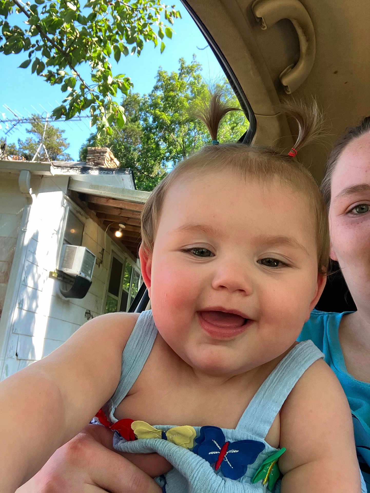 Annabelle is registered to the contest to win money with this photo: baby, child, smiling, pigtails, blue_dress, butterfly, face, person, happy, outdoor, car, window, tree, leaf, sky, adult, hand, clothing, portrait, sunlight