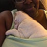puppy, dog, white_dog, sleeping, blanket, wrapped, person, glasses, smiling, selfie, close_up, indoor, cozy, cuddling, pet, human, portrait, warm_lighting, seated, home
