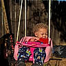 child, toddler, swing, pink_swing, pajama_pants, outdoor, sunny, curly_hair, smile, playful, shoes, rope, wooden_post, fence, backyard, daylight, relaxed, happy, clothing, person