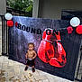 child, toddler, boxing_gloves, banner, balloons, shirtless, shorts, sneakers, outdoor, pavement, celebration, party, fence, curly_hair, confident, decorations, standing, red, black, white