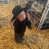 toddler, child, black_hat, wood_shavings, pen, fence, baby_deer, animal, indoor, curious, footwear, clothing, standing, young_child, animal_pen, farm, cute, hat, portrait, daylight