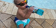 Rylee Rice is registered to the contest to win money with this photo: toddler, child, pool, water, sunglasses, bottle, swim_shorts, barefoot, wet_hair, sitting, outdoor, summer, reflection, drink, sidewalk, tiles, sunny, relaxation, young_child, fun