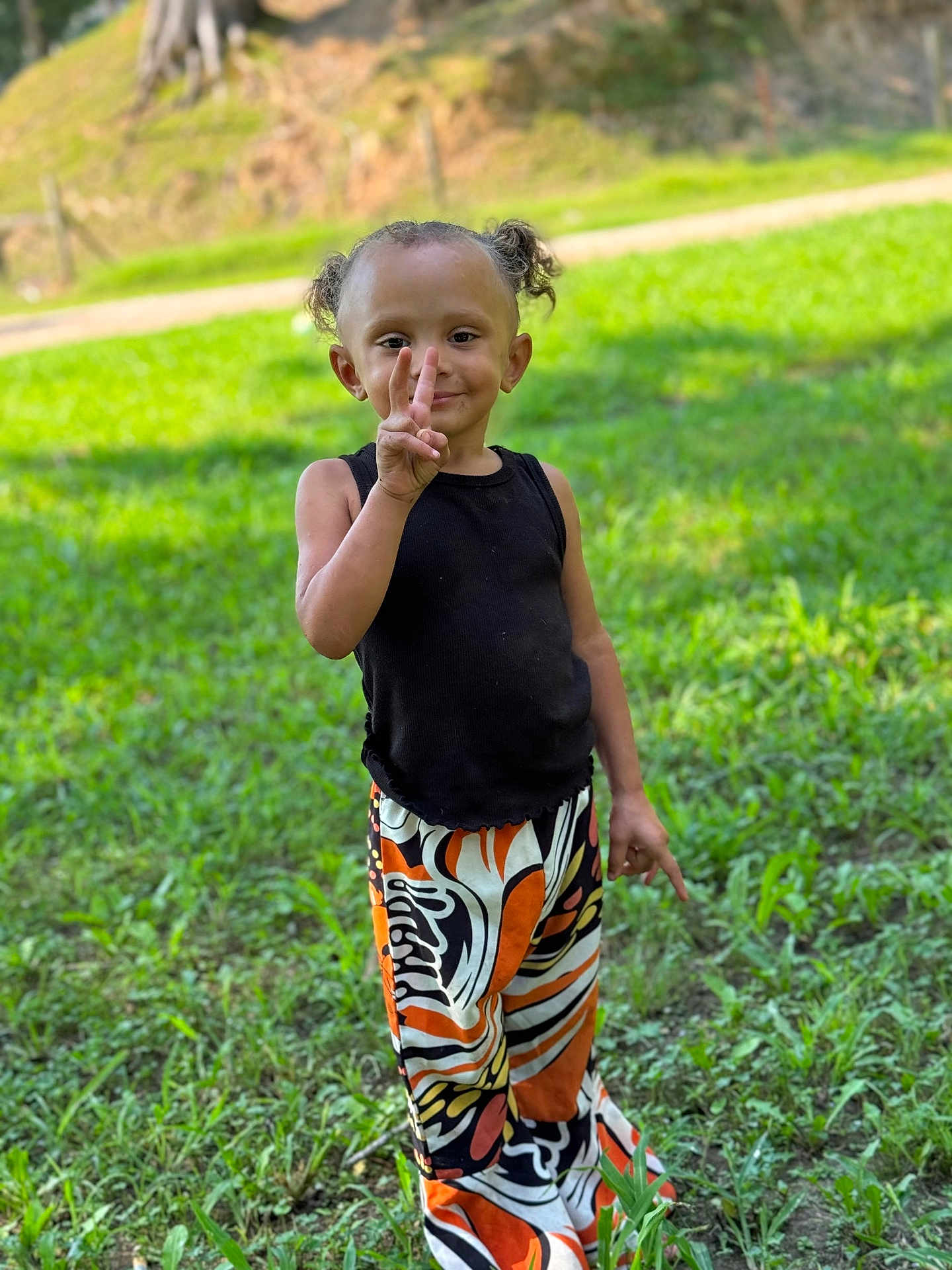 Kinslee joined the competition — help win amazing prizes! child, peace_sign, outdoor, grass, greenery, black_tank_top, colorful_pants, curly_hair, pigtails, smiling, standing, nature, person, playful, happy, casual_clothing, sunlight, daytime, young_child, portrait