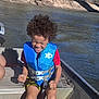 child, boy, life_jacket, boat, river, water, outdoor, summer, sunlight, curly_hair, happy, sitting, clothing, nature, fun, safety, adventure, bridge, daytime, person