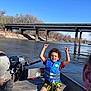 child, boy, life_vest, boat, river, bridge, water, outdoor, nature, trees, sky, summer, smiling, happy, casual_clothing, curly_hair, adventure, recreation, sunny, daytime