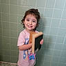 Elora is registered to the contest to win money with this photo: child, smiling, curly_hair, tiled_wall, tiled_floor, bathroom, book, pink_tshirt, cartoon_print, standing, person, indoors, happy, young_child, portrait, casual_clothing, barefoot, holding_object, cute, playful