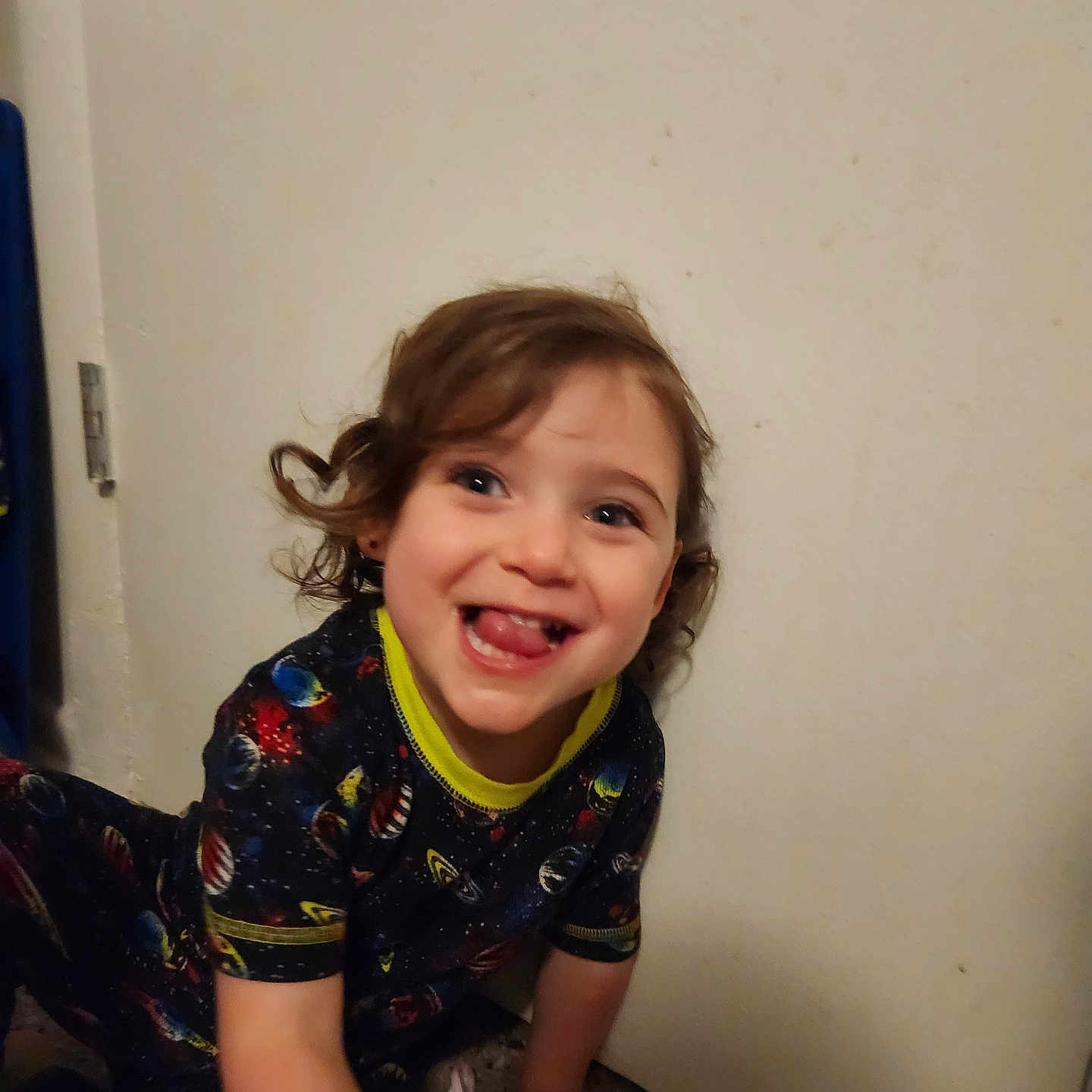 Elora joined the competition — help win amazing prizes! toddler, child, curly_hair, smiling, tongue_out, space_outfit, indoor, floor, wall, playful, happy, person, clothing, young_child, cute, expression, sitting, toy, casual, portrait