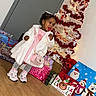 toddler, child, lollipop, white_coat, pink_dress, handbag, christmas_tree, christmas_decorations, gift_bags, holiday, indoor, wooden_floor, smiling, festive, holiday_season, present, cute, shoes, grey_leggings, happy