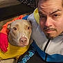man, dog, yellow_sweater, selfie, indoor, brown_dog, facial_hair, braided_hair, colorful_jacket, black_pants, blue_bear_designs, wooden_floor, floral_fabric, close_up, pet, companion, casual_clothing, portrait, warm_lighting, calm_expression