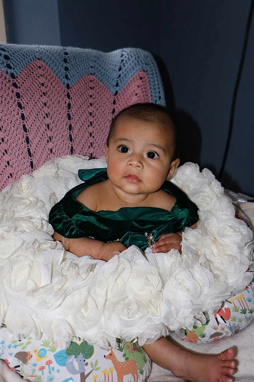 Ailani is registered to the contest to win money with this photo: animal_print, baby, child, colorful_pillow, curious, cute, decor, fabric, face, foot, green_dress, head, indoor, infant, person, portrait, sitting, soft_texture, white_cushion, young_child