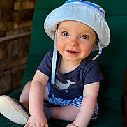 Greyson is registered to the contest to win money with this photo: baby, child, smiling, hat, blue_shirt, striped_shorts, cushion, green_chair, cute, happy, indoor, portrait, infant, hands, feet, clothing, face, person, sitting, sun_hat