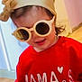 accessory, casual_clothing, child, cute, fashion, floor, hair_bow, headband, home, indoor, person, playful, portrait, red_shirt, smiling, sunglasses, text_on_clothing, toddler, wall, young_child