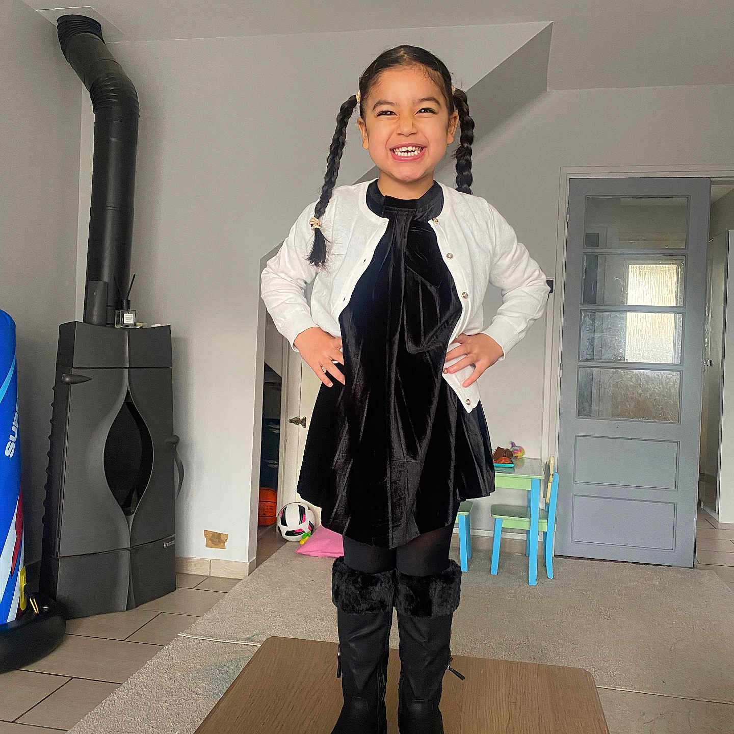 Jane participe au concours pour gagner de l'argent avec cette photo : black_dress, boots, carpet, chair, child, door, floor, girl, happy, home, indoor, kids_table, pigtails, portrait, smiling, soccer_ball, standing, stove, table, white_cardigan
