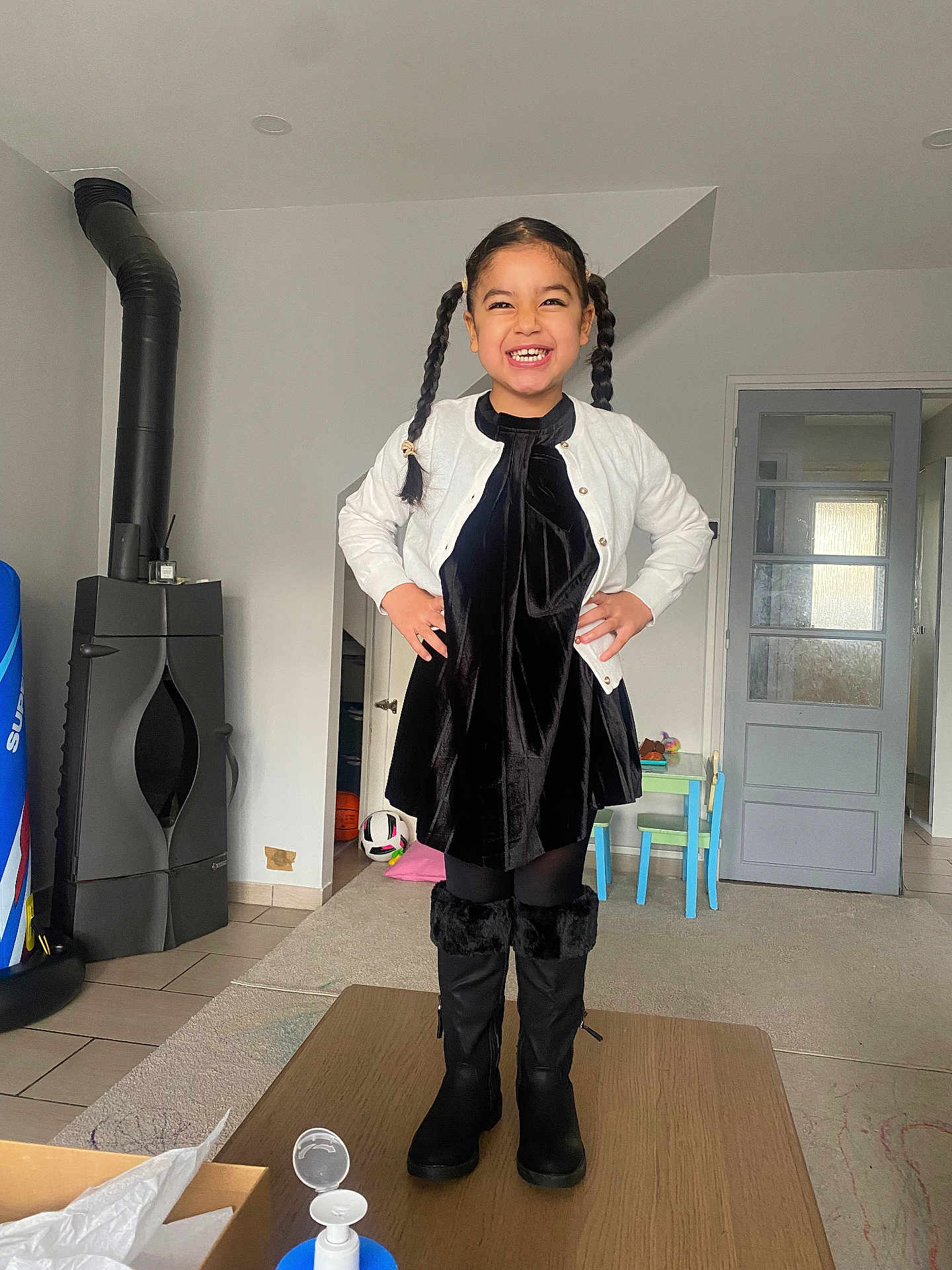Jane participe au concours pour gagner de l'argent avec cette photo : child, girl, smiling, standing, pigtails, black_dress, white_cardigan, boots, indoor, table, carpet, door, kids_table, chair, soccer_ball, floor, stove, home, happy, portrait