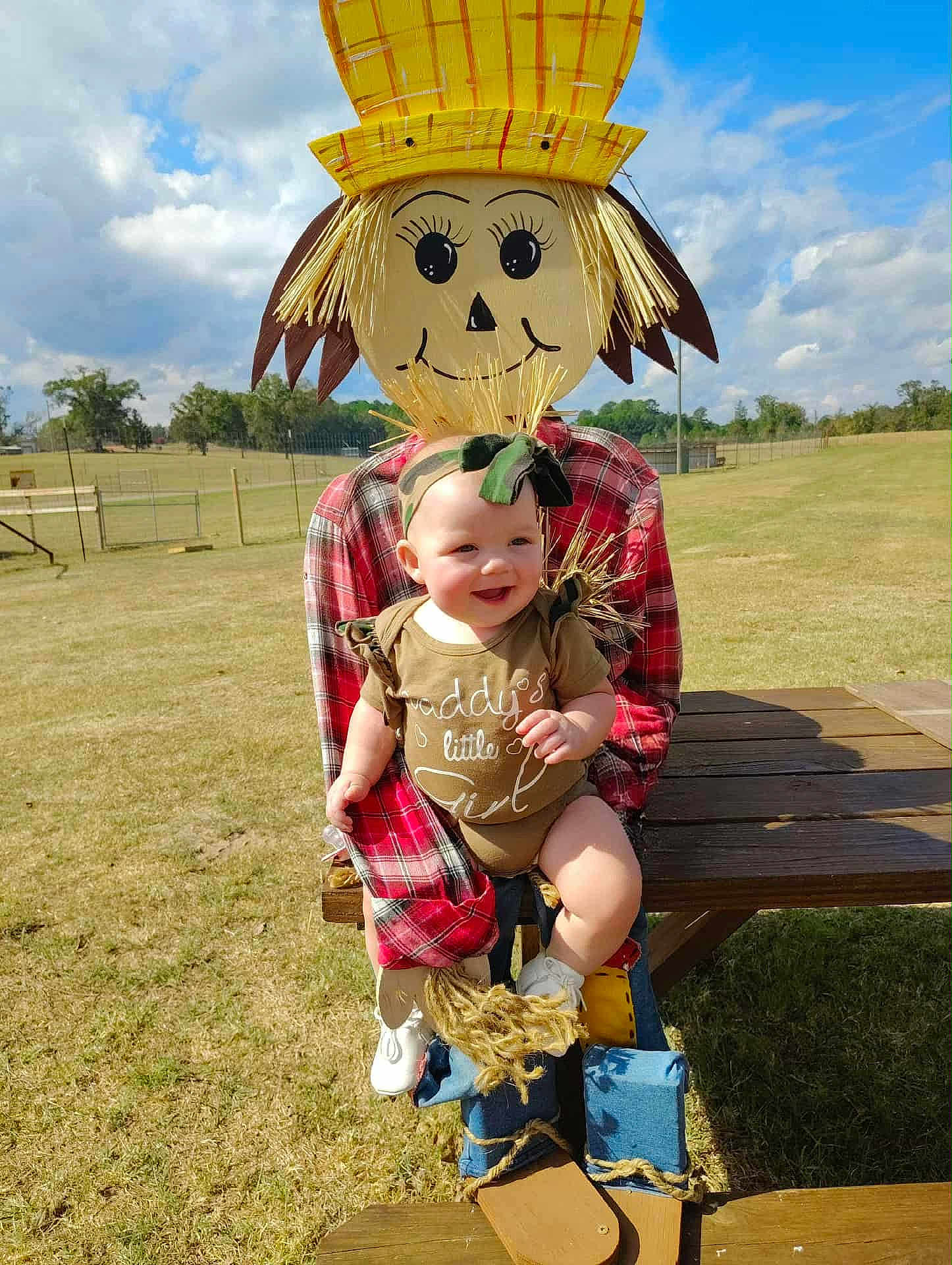 Nellie joined the competition — help win amazing prizes! baby, child, scarecrow, outdoor, grass, blue_sky, clouds, plaid_shirt, headband, smiling, table, wood, field, sunny, person, toy, cute, green, footwear, nature