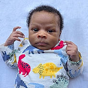 Maro is registered to the contest to win money with this photo: baby, infant, child, dinosaur_print, clothing, onesie, hand, face, eyes, lying_down, blanket, cute, portrait, skin, expression, young, person, indoor, soft, colorful