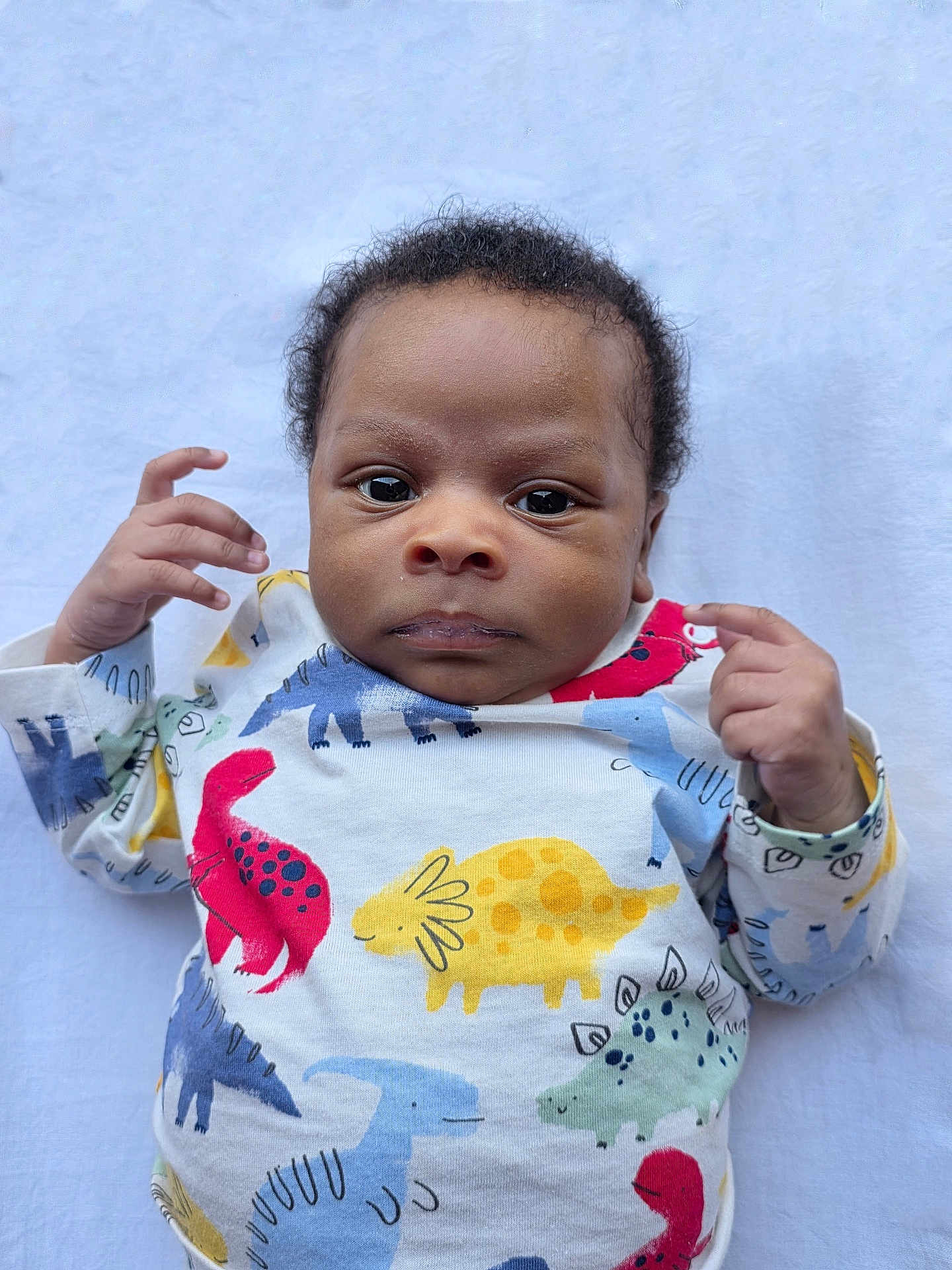 Maro is registered to the contest to win money with this photo: baby, infant, child, dinosaur_print, clothing, onesie, hand, face, eyes, lying_down, blanket, cute, portrait, skin, expression, young, person, indoor, soft, colorful